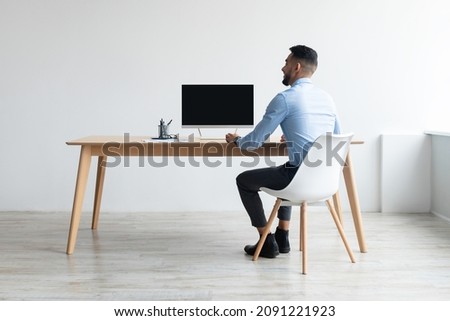 Rear back view portrait of Middle Eastern businessman looking at blank empty computer monitor sitting at desk at workplace, watching webinar or having online web video call, free copy space banner Royalty-Free Stock Photo #2091221923