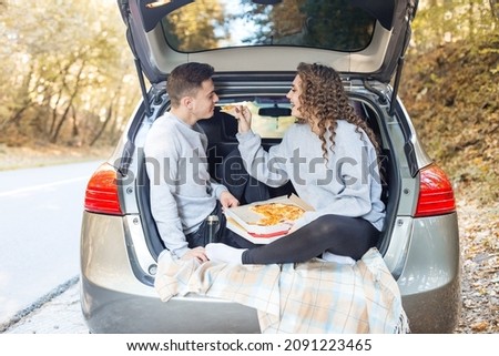 A man and a woman are eating pizza near the car. Picnic in the fall.