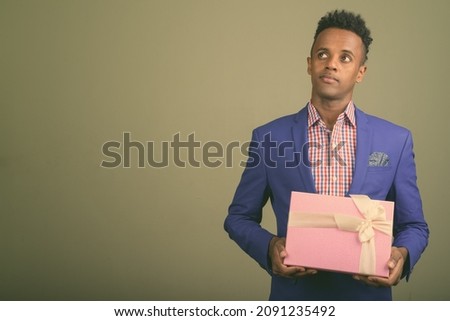 Studio shot of young handsome African businessman wearing suit while holding gift box against colored background