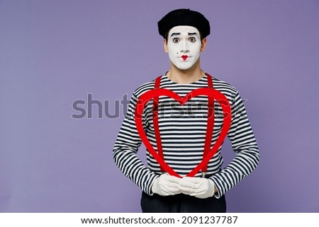Charismatic amazing surprised puzzled fun romantic young mime man with white face mask wears striped shirt beret holding big red heart isolated on plain pastel light violet background studio portrait