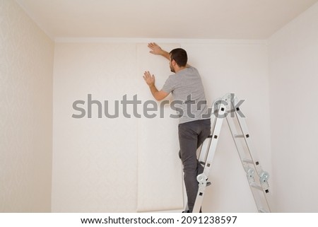 Young adult man standing on metal ladder and applying new wallpaper on white wall in room. Repair work of home. Making interior change. Back view. Royalty-Free Stock Photo #2091238597