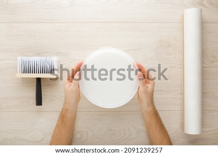 Young adult man hands holding bucket with glue. Roll of wall paper, brush on light wooden table background. Closeup. Point of view shot. Preparing for repair work of home. Ready to glue wallpaper.  Royalty-Free Stock Photo #2091239257