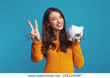 Excited young girl in casual orange sweater holding white piggy bank with lots of money, showing peace gesture over blue background