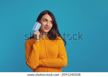 Thoughtful young woman wearing yellow clothes holding plastic credit card near face and looking at free empty space, isolated over blue background  Royalty-Free Stock Photo #2091240508