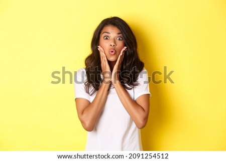 Portrait of attractive african-american teenage girl looking amazed, saying wow and stare at camera excited, standing over white background