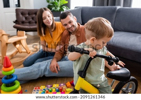 Toddler kid standing near bicycle and blurred parents in living room