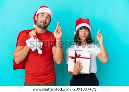 Young couple with christmas hat handing out gifts isolated on blue background with fingers crossing and wishing the best