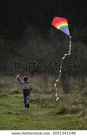 Cute little Caucasian boy runs on green grass in field and launches multicolored striped kite in shape of rhomb with long tail into sky. Actively spend childhood in nature. Lifestyle concept.