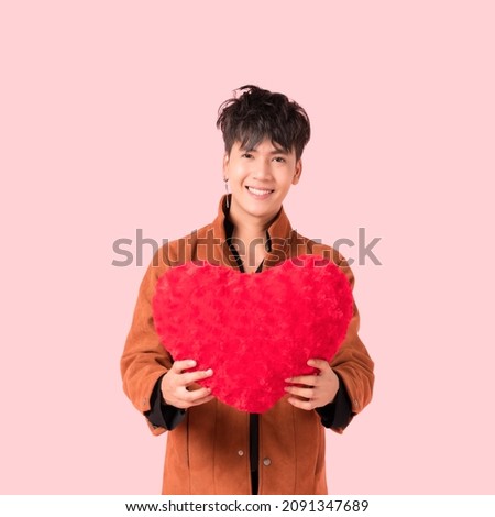 Portrait of happy handsome Asian young man in fashionable clothes standing smiling and holding red heart shape pillow with look at camera in studio isolated on pink background. Valentine's day concept