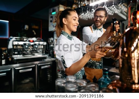 Young happy bartenders pouring beer from a beer tap while working in a pub. Focus is on woman. Royalty-Free Stock Photo #2091391186