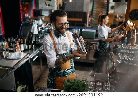Young happy bartender using cocktail shaker while making a drink in a pub. Royalty-Free Stock Photo #2091391192