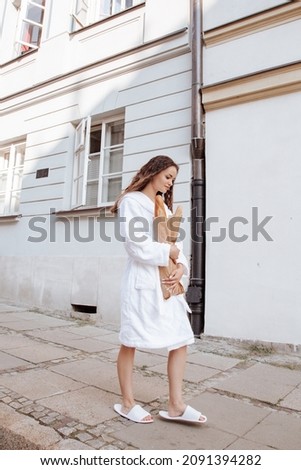 Young woman in white robe looks at street of city. Attractive girl holding baguettes and paper bag with purchases. Young stylish woman buying a french baguette standing on the street in Warsaw city.