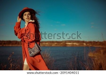 Fashionable woman wearing trendy orange trench coat, hat, holding zebra print faux leather handbag, posing in nature. Outdoor autumn fashion portrait. Copy, empty space for text