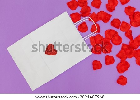 On a purple background is a white paper shopping bag on which is a red glitter heart. From the bag come out red petals, which are scattered.