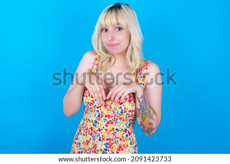 Caucasian girl wearing floral dress isolated over blue background makes bunny paws and looks with innocent expression plays with her little kid
