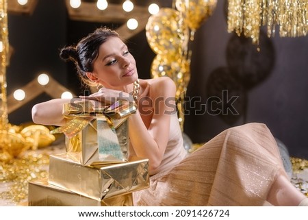 Beautiful young woman on a golden background with gifts. Stylish foil sequins and balloons. Gold and black colors. Trendy stylish girl. Disco nightlife. Birthday anniversary celebration. New Year 