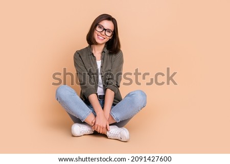 Photo of cheerful lovely lady sit floor cuddle legs beaming smile wear specs khaki shirt isolated beige color background