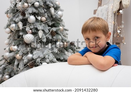 A little blond boy in Christmas pajamas stands by a Christmas tree and leans on a white leather chair, looking straight ahead. Christmas concept, holiday anticipation.