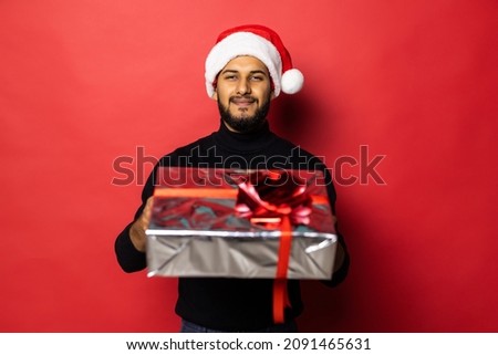 Young indian man in Santa hat holding Christmas gift standing on red background