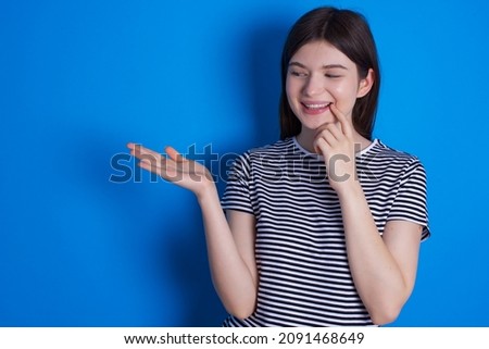 Positive Caucasian woman wearing striped T-shirt isolated over blue background advert promo touch finger teeth