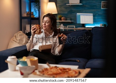 Smiling woman sitting on couch looking at tasty takeaway burger holding beer bottle in front of table with pizza and takeaway junk food menu. Happy person eating tasty burger and delivery dinner.