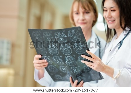 Doctor in uniform, a stethoscope, and a female assistant stand together in a hospital corridor. Consulting or checking the X-ray result sheet. Asian and European women.
