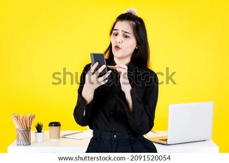 Studio shot of Asian confused amazed doubt thoughtful female businesswoman employee in black outfit sitting on working desk checking news on e-mail from smartphone at office on yellow background.