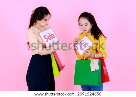 Portrait studio shot Asian young happy female shopper wearing casual outfit carrying colorful shopping bags holding showing sale discount banner paper on pink background.