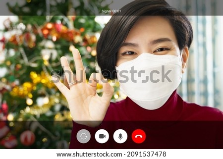 Web cam video call shot mock up. A beautiful asian woman in red with medical face mask happy smiling and do okay hand sign to her friends and family through online chat application on Christmas eve.