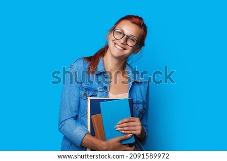 Portrait of a caucasian student girl wearing eyeglasses smiling and holding exercise books isolated over blue background