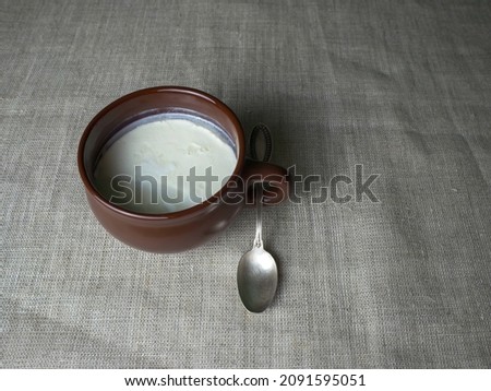 Plain white homemade yogurt in a brown clay cup, next to it is a silver spoon on a gray linen tablecloth. Minimalistic food background