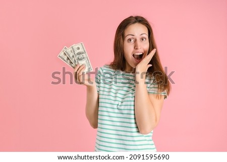 A brunette woman holds a bundle of dollar bills in her hand and covers her mouth with her hand in surprise. Pink background