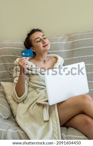 beautiful young girl in home clothes with a card in her hands and a laptop on her knees shopping online. The concept of shopping during a pandemic