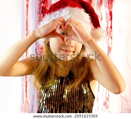 a cute girl with blond hair in a Santa Claus hat stands against the background of shiny decorations on New Years night and shows a heart out of her hands. Portrait of a little girl at a new year party