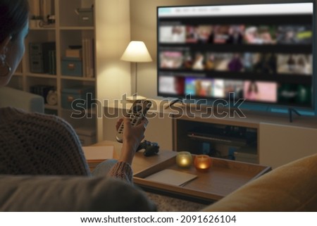 Woman relaxing on the couch, she is using the remote control and choosing a TV show or movie on the television menu Royalty-Free Stock Photo #2091626104