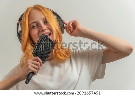 teen girl with red hair in a white t-shirt on a light background listening to music with headphones and singing.