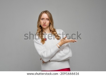 Disappointed blonde young woman pointing hand right, looking upset and frowning skeptical, showing at promo offer, stands in white sweater against gray background