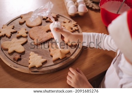 Little girl in santas hat decorates gingerbread using white glaze. Christmas and New Year traditions concept. Christmas bakery. Happy hollidays