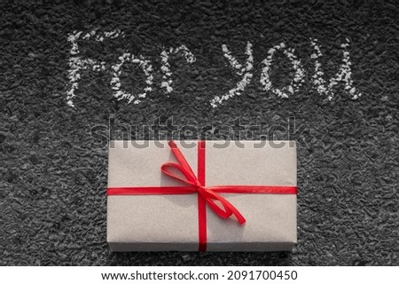 a gift wrapped in craft paper tied with a red ribbon lies on the gray asphalt. Above written in chalk for you