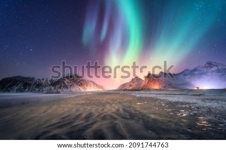Aurora borealis above the snowy mountain and sandy beach in winter. Northern lights in Lofoten islands, Norway. Starry sky with polar lights. Night landscape with aurora, frozen sea coast, city lights Royalty-Free Stock Photo #2091744763