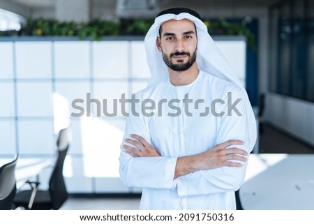 Handsome man with dish dasha working in his business office of Dubai. Portraits of a successful businessman in traditional emirates white dress. Concept about middle eastern cultures Royalty-Free Stock Photo #2091750316