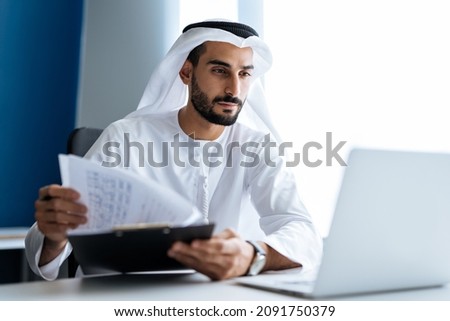 Handsome man with dish dasha working in his business office of Dubai. Portraits of a successful businessman in traditional emirates white dress. Concept about middle eastern cultures Royalty-Free Stock Photo #2091750379