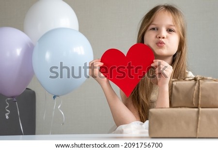 Cute little girl holding big red paper heart and blows a kiss. Child surrounded by zero waste gifts and air balloons. Valentines Day, love, mothers Day, romantic concept. 