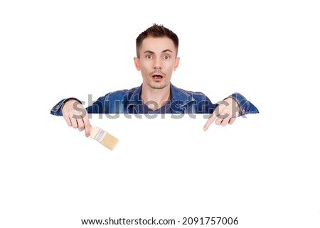 Young painter in denim jacket with surprised expression holds paint brush in hand and points to empty space for ad text. Copy space.