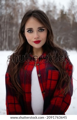 Charming female with red lips and in checkered shirt touching hair while standing in snowy winter woods and looking at camera