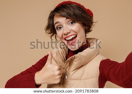 Close up young smiling happy woman in red turtleneck vest beret doing selfie shot pov on mobile phone show thumb up gesture isolated on plain pastel beige background studio. People lifestyle concept