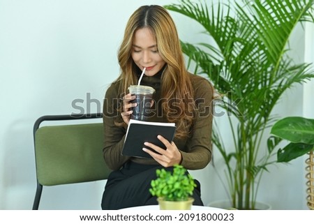Photo of an attractive woman drinking an iced coffee and reading a book in the comfortable living room.