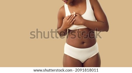 Beautiful plus size young black woman in underwear touches her chest when she feels strong pain standing isolated on beige color banner background. Breast cancer and heart attack awareness concept Royalty-Free Stock Photo #2091818011