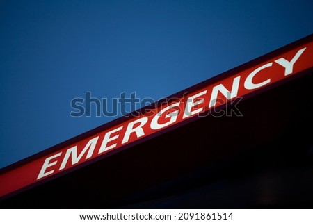 Selective focus illuminated sign that spells EMERGENCY diagonally across the frame, using white letters on a red background, with a deep dark blue sky above and black background below