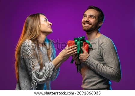 Portrait of happy young couple, woman giving to man his holiday present isolated over purple background in neon lights. Concept of love, relationship, facial expression, emotions, feelings and ad